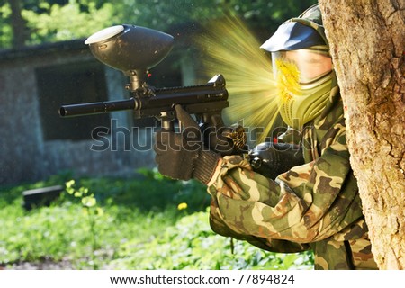 paintball sport player wearing protective mask aiming gun from shelter under gunfire attack with paint splash