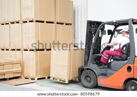 warehouse worker driver in uniform loading cardboard boxes by forklift stacker loader