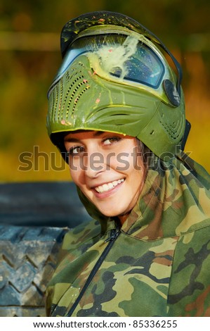 Happy paintball sport player girl in protective camouflage uniform and mask with marker gun outdoors