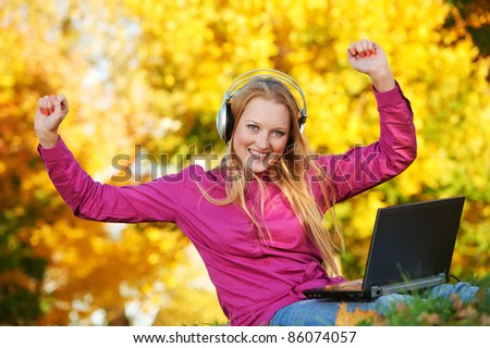 Smiling young attractive woman with headphones and laptop computer in park at fall outdoors