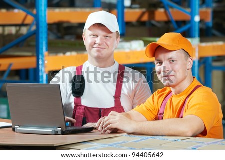 two young handsome workers man in uniform in front of warehouse rack arrangement stillages using notebook computer