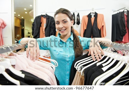 Young smiling woman seller assistant with hands on clothing hangers at shopping store