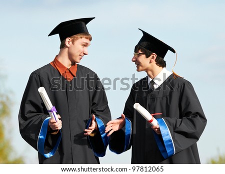 two male graduate students in gown holding diploma over blue sky