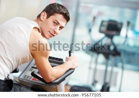 smiling fitness man at legs muscles exercises with bicycle training machine station in gym