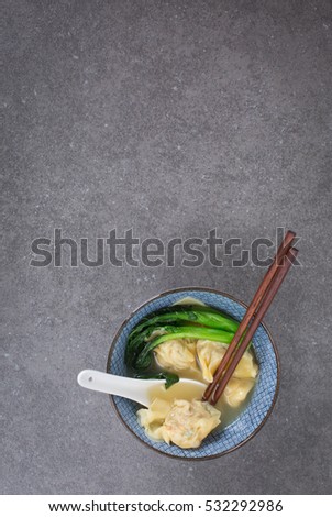Wonton soup in a blue ceramic bowl served wth Gai lan. A ceramic spoon and a pair of bamboo chopsticks. Gray dark stone background.