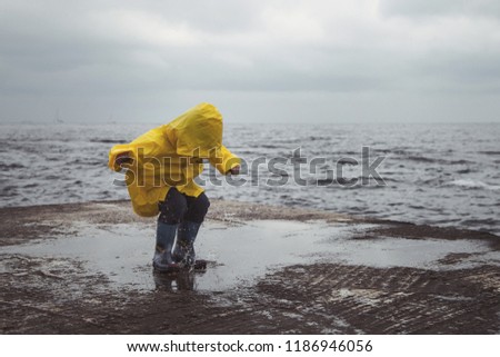 A boy in a yellow raincoat jumps on puddles on the beach.