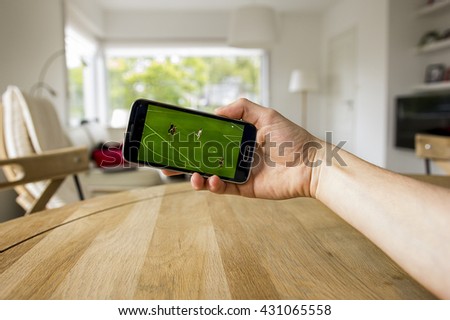 A male hand holding a mobile phone which displays a soccer match on the touch screen. An image on an interior background.
