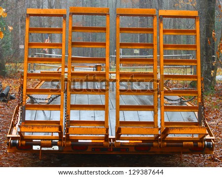 An old large orange wooden floor trailer with loading ramps for hauling tractors and equipment from place to place parked in the woods on a foggy day