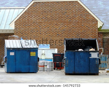 A couple of large blue trash dumpsters with a trash can, some Kegs and an Ice locker behind a business.