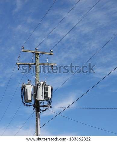 A power utility pole with three transformers lines and insulators against a blue cloud filled sky