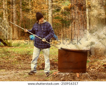 A middle aged man with a mullet in a hat working outside burning leaves and debris inside a fire ring around an old tree stump with a pitch fork using filters layers and textures for a grunge look.