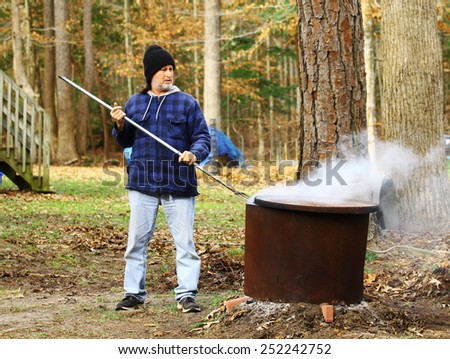 A middle aged man with a mullet wearing a hat working outside during the winter burning leaves and debris inside of a fire ring around an old tree stump with a pitch fork