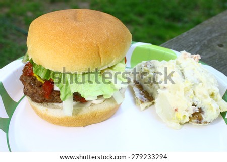 A fresh made hamburger and potatoe salad on a paper plate on an old  wooden picnic table outside during the summer