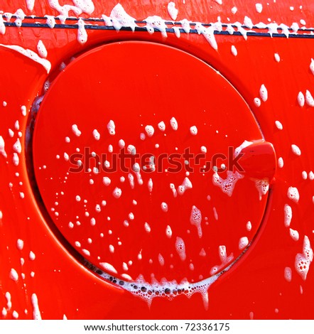 A fuel door on the side of a red car that is being cleaned and washed after all of the bad weather with soap bubbles and water running down the side and room for your text