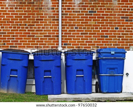 Four blue plastic trash cans lined up along a brick wall on the grass and a concrete platform with room for your text.