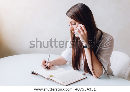 A young beautiful receptionist and personal assistant is very serious. She is making a phone call and taking notices as a business woman
