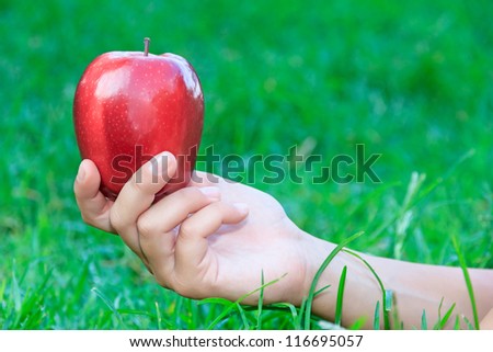 Hand offering a red apple against the grass as a background, focus on the apple