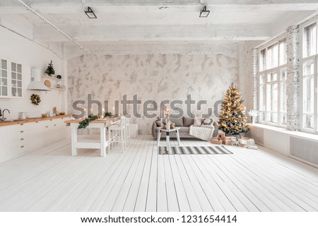 Loft style Apartment, large spacious living room with dining table and kitchen. Room with Christmas tree. Comfortable sofa, high large Windows. Light white brick wall.
