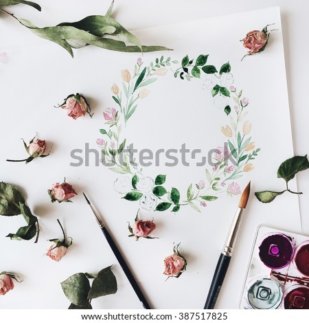 Workspace. Wreath frame with flowers and leaves painted with watercolor, paintbrush and pink roses isolated on white background. Overhead view. Flat lay, top view
