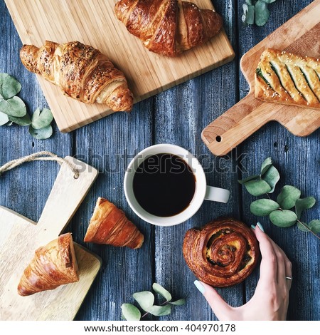 Breakfast with croissants, leaves, cutting board and black coffee composition with girl hand on wooden retro background. Flat lay, top view