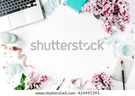 Workspace with paintbrush, laptop, lilac flowers bouquet, spool with beige and blue ribbon, mint diary on white background. Flat lay, top view