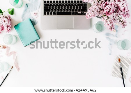 Workspace with paintbrush, laptop, lilac flowers bouquet, spool with beige and blue ribbon, mint diary on white background. Flat lay, top view