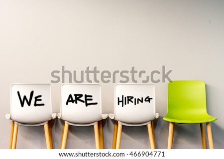Job recruiting advertisement represented by \'WE ARE HIRING\' texts on the chairs or wall. One chair is colored differently to represent the hiring position to be recruited and filled.