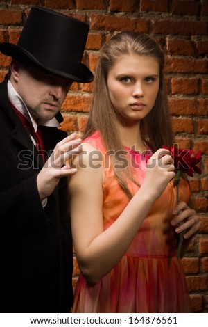 Vampire and his victim. Man in the black coat, top hat and in a red tie and beautiful girl with rose