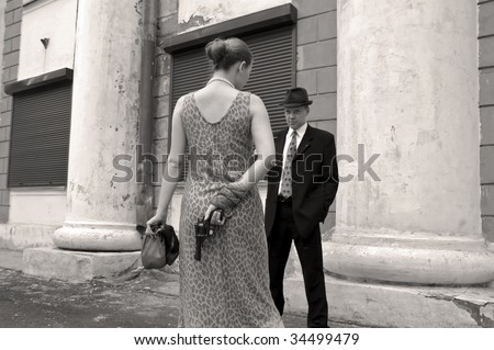 Beautiful stylish girl with a revolver before the man in a business suit. Grayscale image