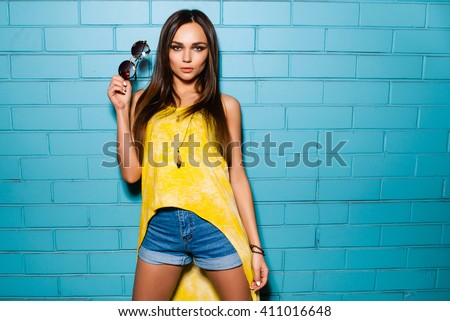 Beautiful young sexy hipster girl posing and smiling near urban blue wall background in yellow dress, shorts, shirt, sneakers, sunglasses.