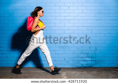 Beautiful young sexy hipster girl goes near urban blue wall background in yellow swimsuit, white jumpsuit, sneakers, sunglasses, pink backpack.