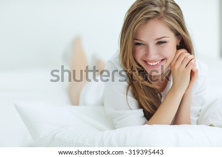 A woman lying at the end of the bed underneath the quilt and smiling, with her head resting upon her hand with the other in her hair.