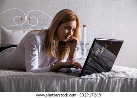 A smiling woman lying down the bed in front of her laptop with her legs raised slightly