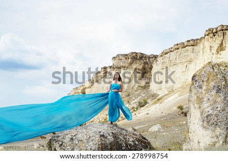 Young beautiful alone woman in dark cyan dress stands on the rock with mountains at background