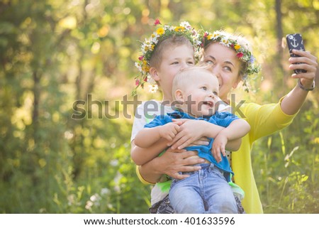 Portrait of adorable kids with their young mother taking selfie in the sunny park. Happy family in flower wreaths making photo with smartphone outdoors on a summer day. Boys and mom smiling.