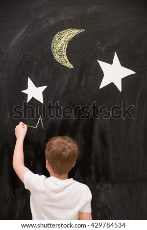 Back view on cute kid boy drawing stars with chalk on a black board with white stars and moon. Child writing on wall in the classroom. Creativity, activities for children indoors. Dreaming about space