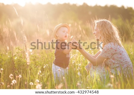 Cute little toddler boy in a straw hat holding his mother\'s hand and blowing dandelion. Adorable child walking with his mom in park on sunny summer day. Family on sunset. Childhood, motherhood concept