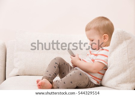 Adorable toddler boy sitting on the sofa in the living room and playing with smartphone. Child learning how to use smartphone. Boy texting on the phone. - technology and lifestyle concept