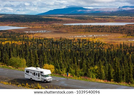 RV, motorhome, caravan parking next to a road in a parking lot in Alaska with spectacular, beautfiul background with lakes and conifer forest, blue sky and clouds
