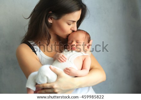 Mother kissing newborn son at grey background. Portrait of woman and little baby