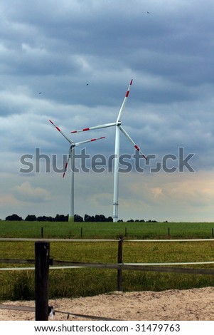On this picture you can see two wind turbines which generate energie. In the background you can see a cloudy sky, and in the front there is sand and gree grass.
