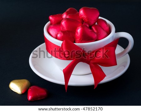 White cup with saucer with red and gold chocolate hearts and red ribbon. Photo on a black background