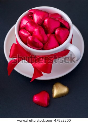 White cup with saucer with red and gold chocolate hearts and red ribbon. Photo on a black background