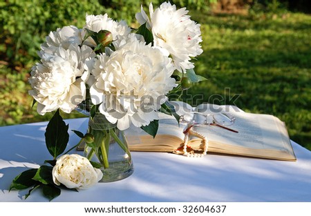A bouquet of white peony flowers in a glass vase on a garden table in summer evening with an open book, a string of pearls and eyeglasses on blur background