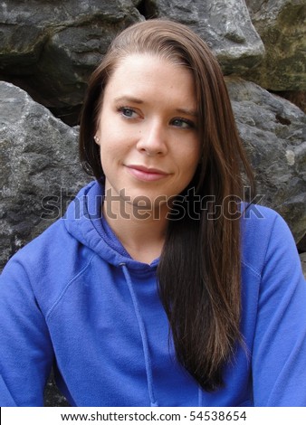 Young woman, blue eyes, brunette, with a slight smile wearing a blue shirt leaning against a large boulder rock wall.