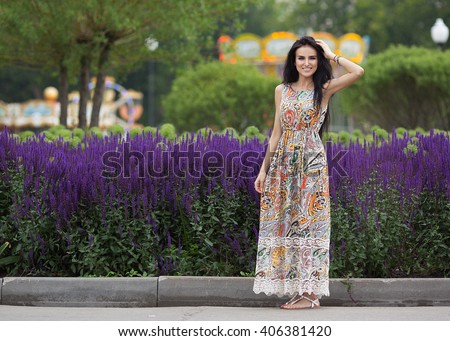 Beautiful young brunette woman in a summer dress standing on the street. The flowers on the background. Summer shot.