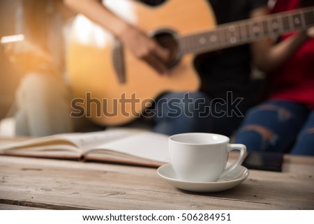 White coffee cup placed on the table, many women are singing and playing the guitar in party.