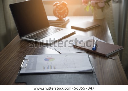 A wooden desk with a laptop, notebook,clock,pen and annual reports in morning,working space.