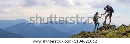 Young tourists with backpacks, athletic boy helps slim girl to clime rocky mountain top against bright summer sky and mountain range background. Tourism, traveling and healthy lifestyle concept.