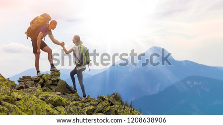 Young tourists with backpacks, athletic boy helps slim girl to clime rocky mountain top against bright summer sky and mountain range background. Tourism, traveling and healthy lifestyle concept.